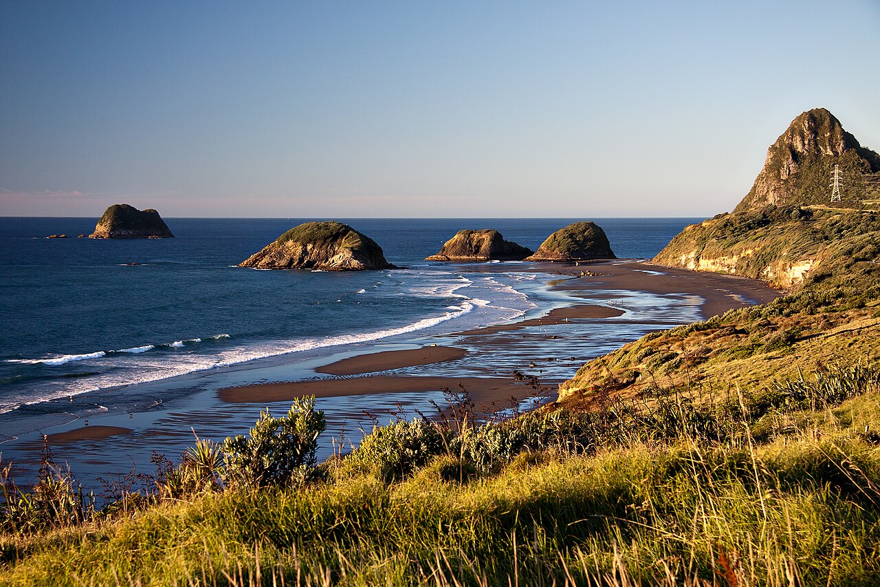 Back Beach below a green slope, with Paritūtū on the right and offshore islands to the left in evening light.