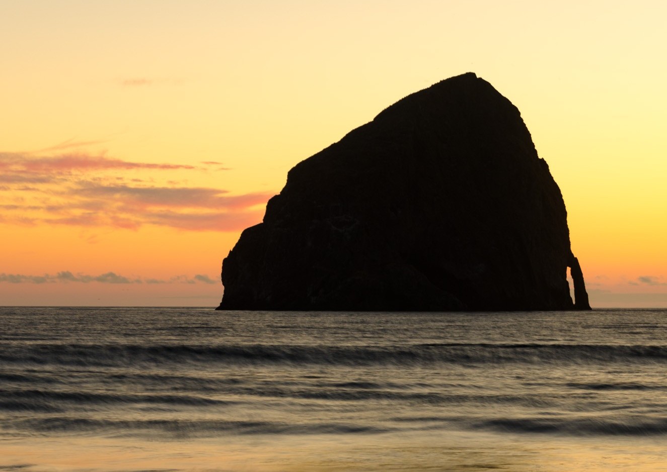 Pacific City's offshore Haystack Rock silhouetted against a sunset, with waves rolling in below it.