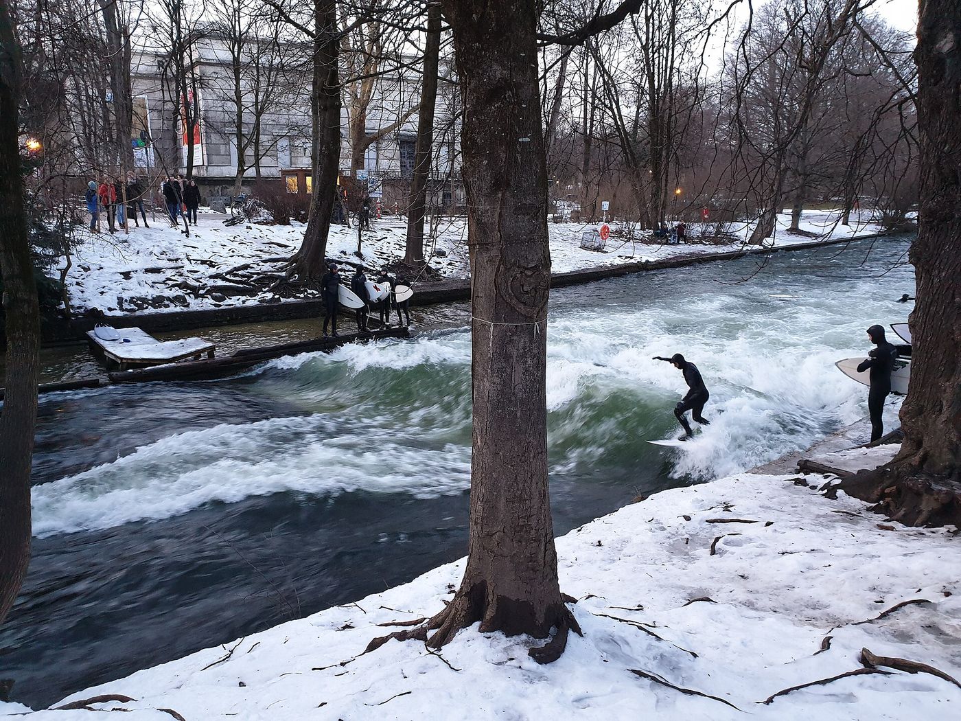 A surfer riding the Eisbach wave in Munich, with snow on the banks and other surfers waiting beside the channel.