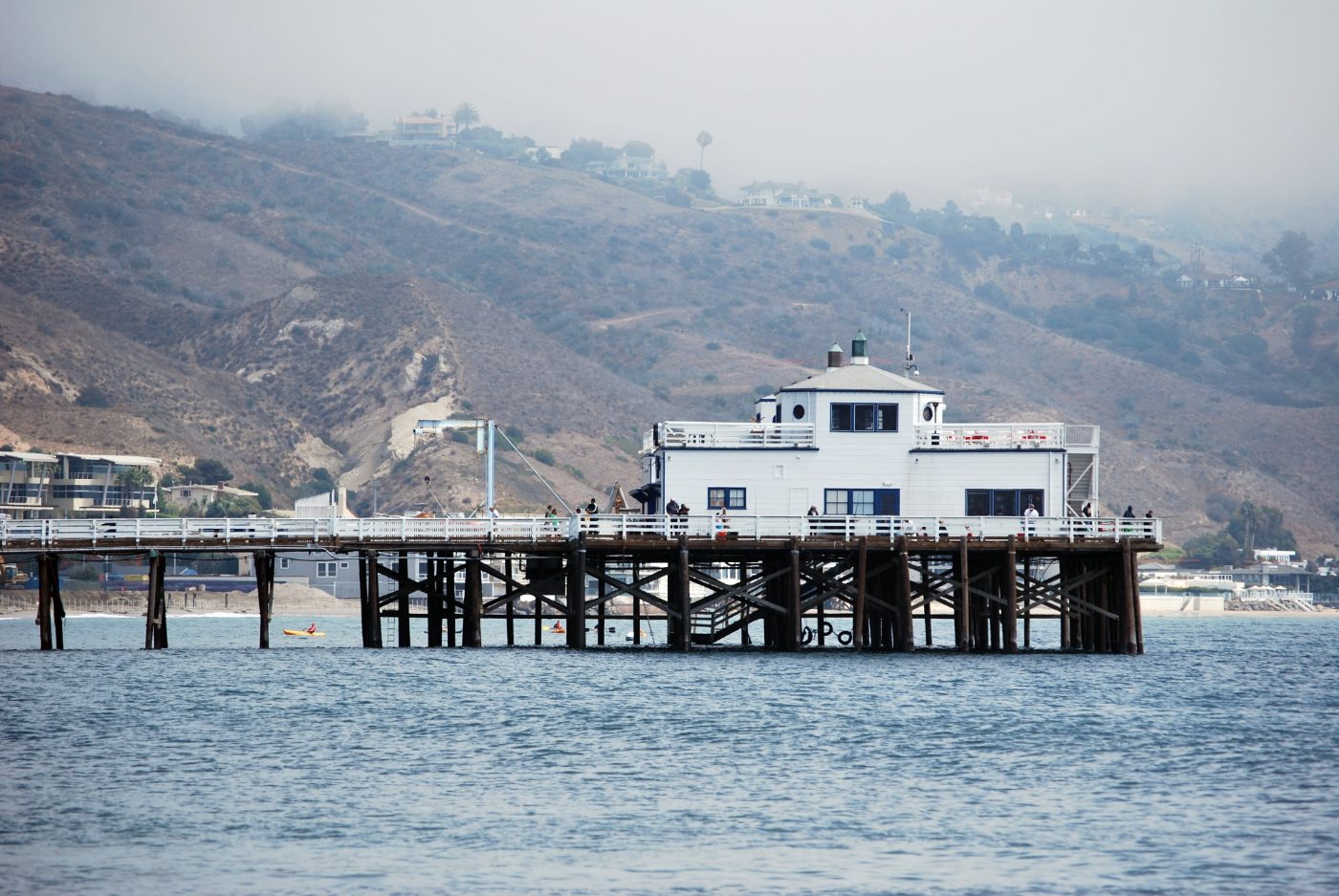 Malibu Pier's white building above the water, with misty coastal hills behind it.