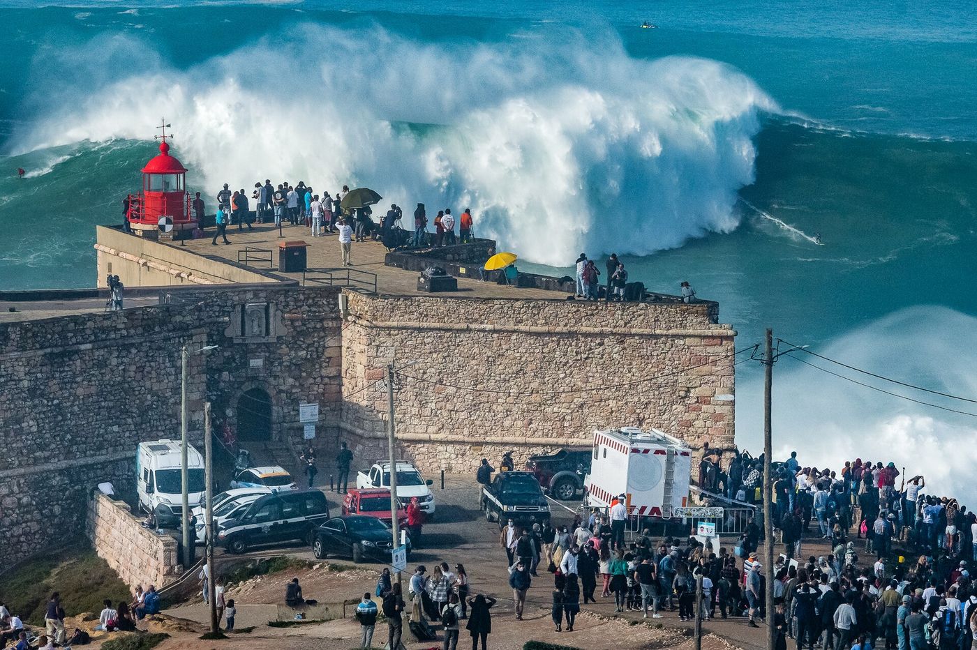 Forte de São Miguel Arcanjo and its red light, with spectators on the roof and a breaking wave behind it at Nazaré.