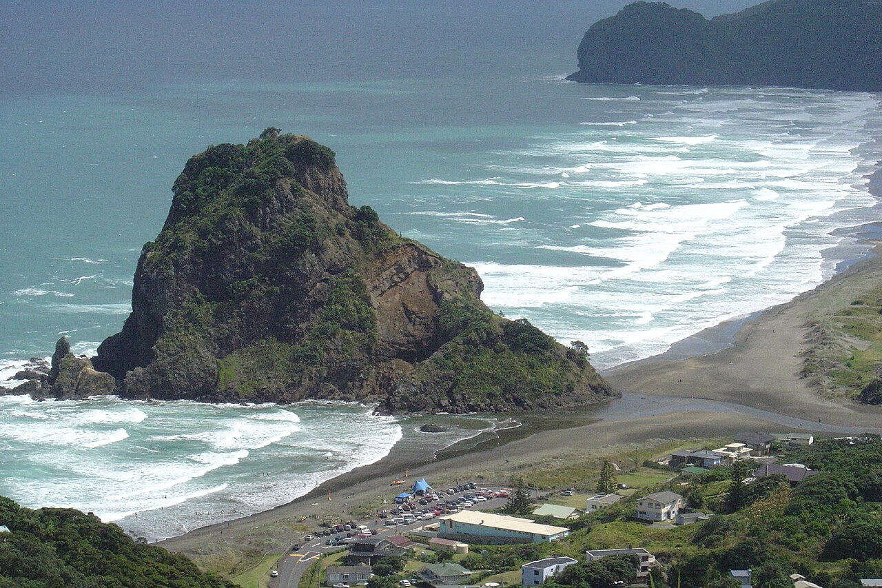 Lion Rock beside Piha Beach, with surf, green slopes and houses in an elevated view.