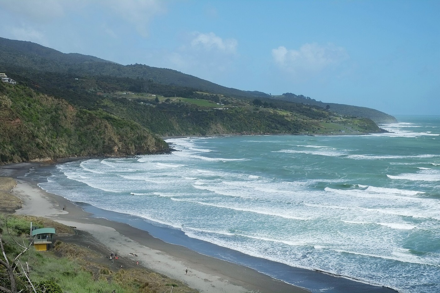 Looking west along the Raglan coast towards Manu Bay, with surf, a beach hut and green hills.