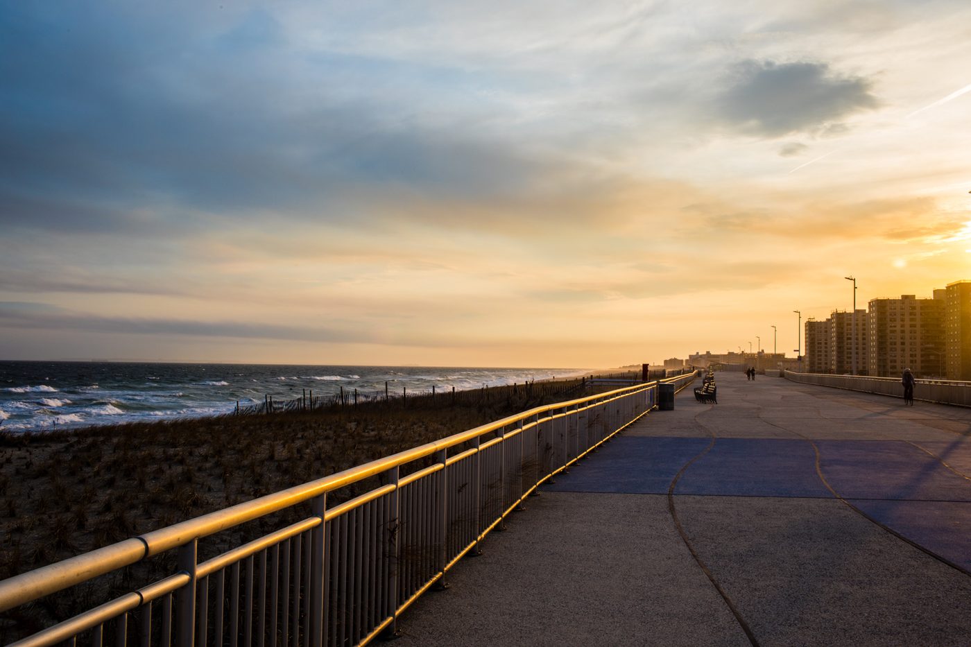 Rockaway's boardwalk at sunset, with the ocean and dune fencing on one side and apartment buildings on the other.