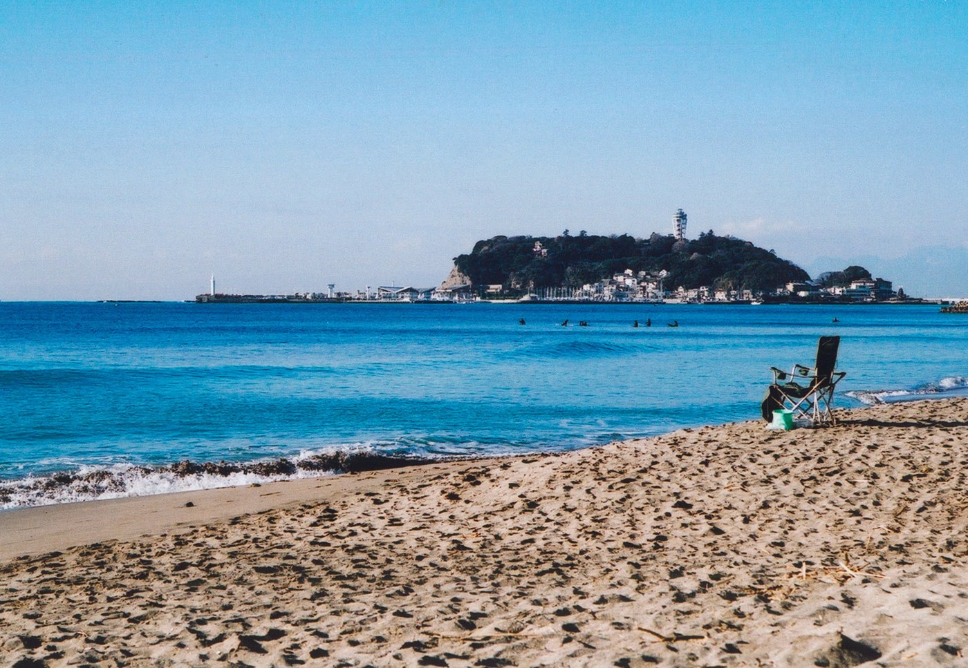Shonan beach with a folding chair on the sand, surfers in the water and Enoshima beyond.