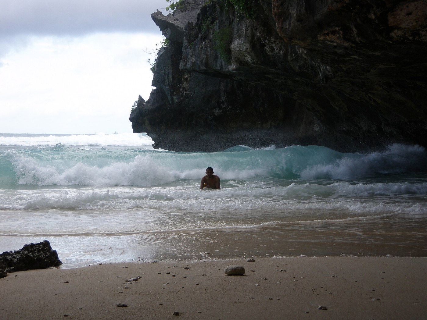 Waves beside a high rock wall at Suluban Beach, with a person in the water and sand in the foreground.