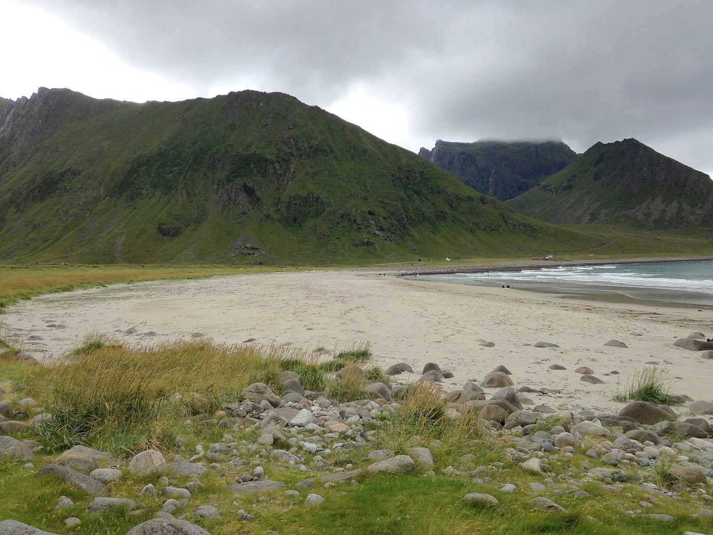 The beach at Unstad in Lofoten, with rounded stones and grass in the foreground and steep green slopes behind.