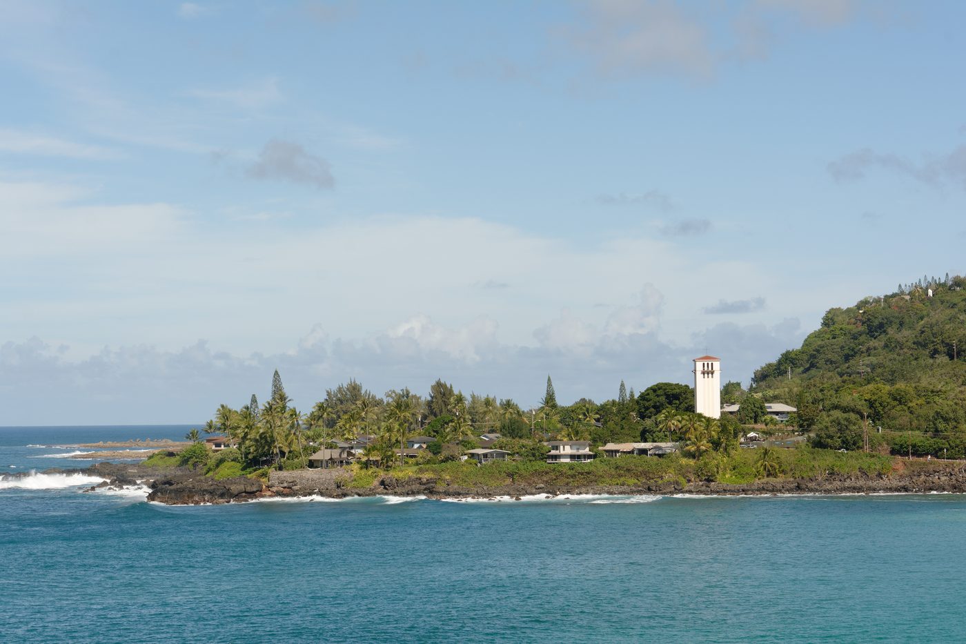 View across Waimea Bay towards a rocky, tree-covered shore and a pale church tower.