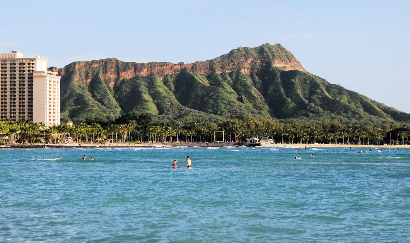 Diamond Head above the Waikīkī shoreline, with palms, buildings and people in the water.